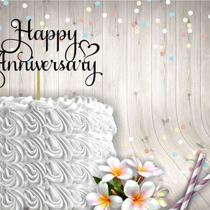 May include: White cake with white frosting and a black cake topper that says "Happy Anniversary". The cake is on a wooden background with colourful confetti.