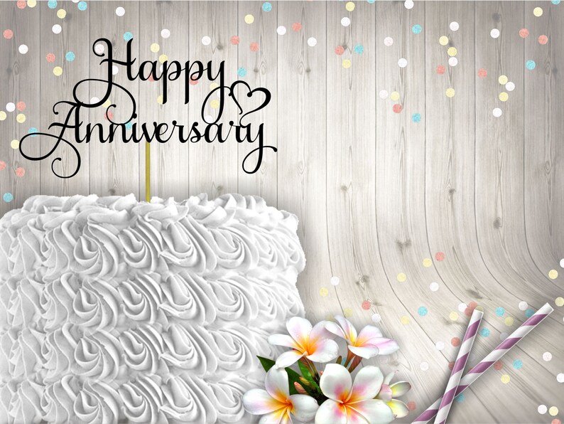 May include: White cake with white frosting and a black cake topper that says "Happy Anniversary". The cake is on a wooden background with colourful confetti.