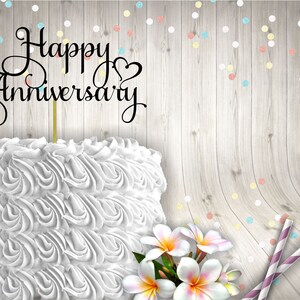 May include: White cake with white frosting and a black cake topper that says "Happy Anniversary". The cake is on a wooden background with colourful confetti.