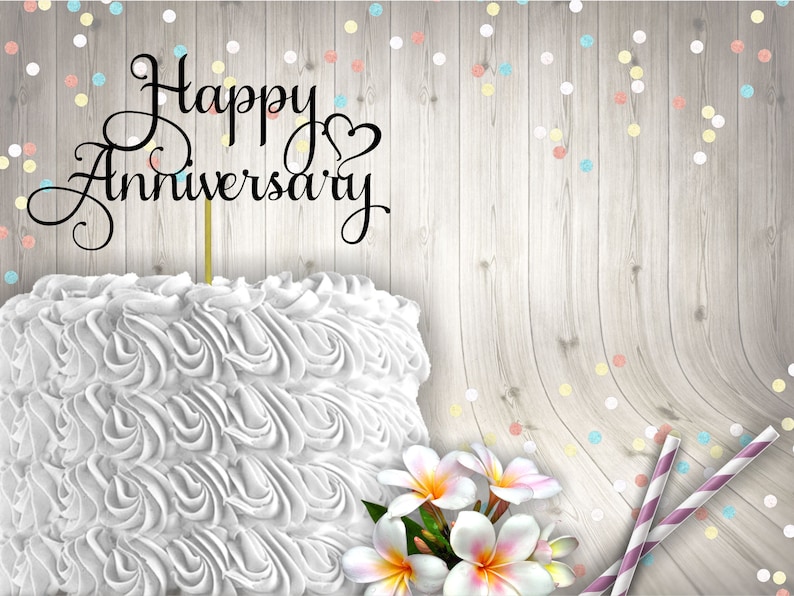 May include: White cake with white frosting and a black cake topper that says "Happy Anniversary". The cake is on a wooden background with colourful confetti.