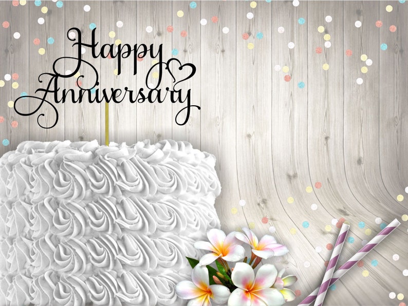 May include: White cake with white frosting and a black cake topper that says "Happy Anniversary" with a heart. The cake is on a wood background with colourful confetti.