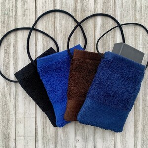 以下が含まれることがあります： 黒、青、茶色、2色の青のテリークロス製の吊り下げ式石鹸ポーチが5つあります。ポーチには黒い紐が付いており、白い木製の背景に吊り下げられています。ポーチの1つには石鹸が入っています。