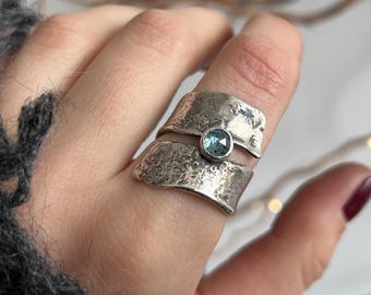 May include: A silver ring with a textured band design and a small, round, light blue gemstone. The ring is worn on a finger, with a gray knitted sleeve in the background. The ring's design is modern and unique.