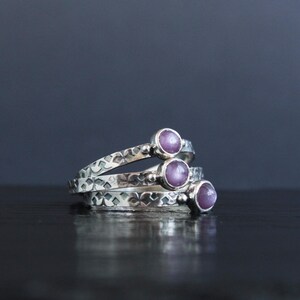 May include: Three silver rings stacked on top of each other, each with a small, round, purple gemstone. The rings have a textured, patterned design. The background is a dark, solid color.