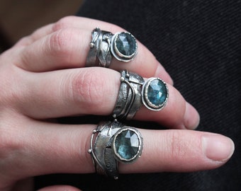 May include: Three silver rings with blue gemstones set in the center of each ring. The rings are stacked on a finger.