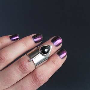 May include: A silver ring with a black gemstone set in a pointed design. The ring is worn on the middle finger of a hand with purple glitter nail polish.