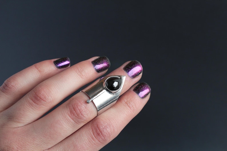 May include: A silver ring with a black gemstone set in a pointed design. The ring is worn on the middle finger of a hand with purple glitter nail polish.