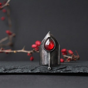 May include: A silver ring with a pointed top and a red gemstone in the center. The ring is on a black slate surface.