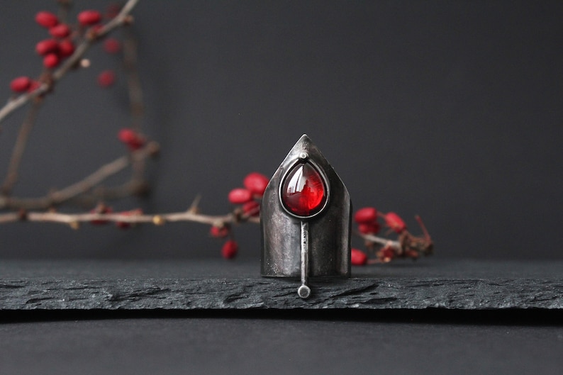 May include: A silver ring with a pointed top and a red gemstone in the center. The ring is on a black slate surface.