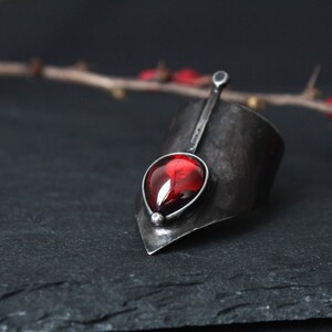 May include: A silver ring with a pointed design and a red gemstone. The ring is set on a dark gray surface.