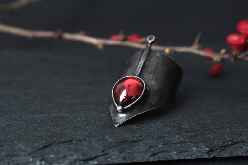 May include: A silver ring with a pointed design and a red gemstone. The ring is set on a dark gray surface.