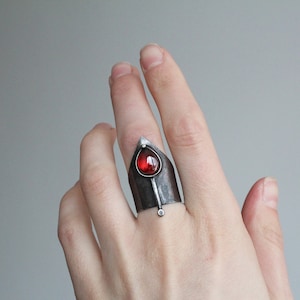 May include: A silver ring with a red gemstone set in a pointed, triangular design. The ring is worn on the middle finger of a hand.