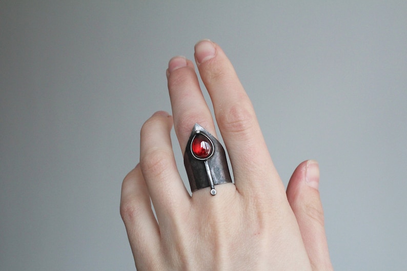 May include: A silver ring with a red gemstone set in a pointed, triangular design. The ring is worn on the middle finger of a hand.