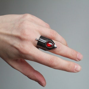 May include: A silver ring with a red gemstone and a pointed tip. The ring is worn on a finger.