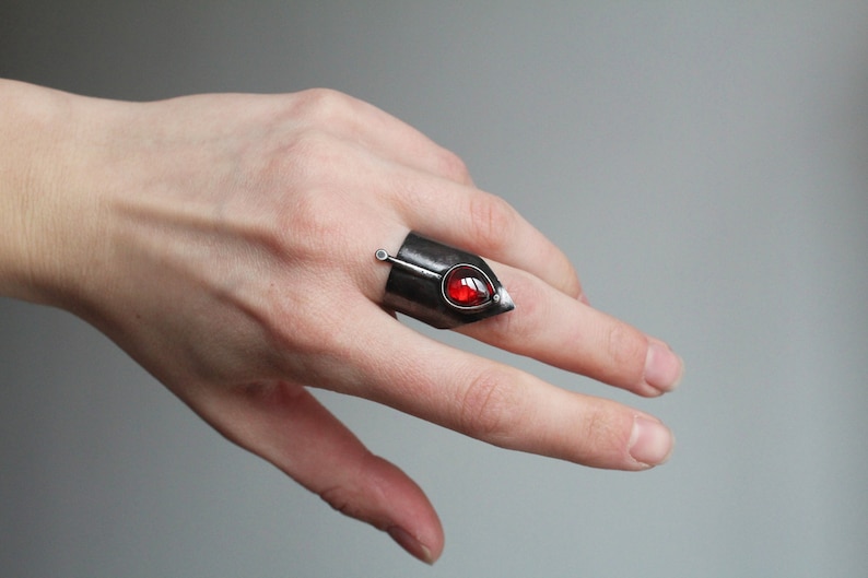 May include: A silver ring with a red gemstone and a pointed tip. The ring is worn on a finger.