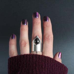 May include: A silver ring with a black gemstone set in a pointed design. The ring is worn on the middle finger of a hand with purple glitter nail polish.