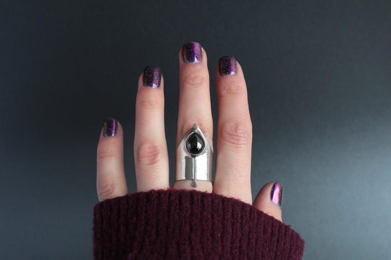 May include: A silver ring with a black gemstone set in a pointed design. The ring is worn on the middle finger of a hand with purple glitter nail polish.