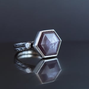 Op de afbeelding: Zilveren ring met een gefacetteerde, zeshoekige, donkerrode edelsteen. De ring heeft een getextureerde band met een kleine, ronde accent. De edelsteen reflecteert licht, tegen een donkere achtergrond.