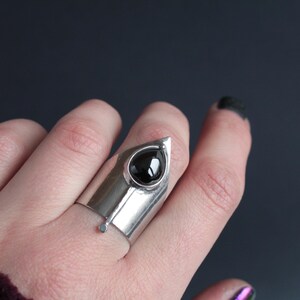May include: A silver ring with a black gemstone set in a pointed, triangular design. The ring is worn on a finger.