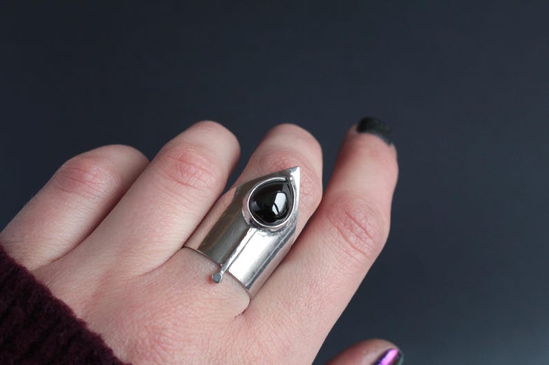 May include: A silver ring with a black gemstone set in a pointed, triangular design. The ring is worn on a finger.