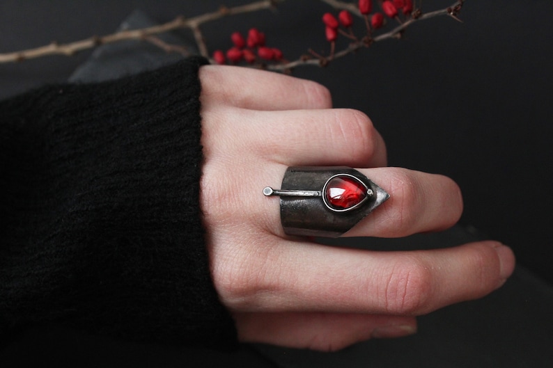 May include: A silver ring with a pointed design and a red gemstone. The ring is worn on a finger.