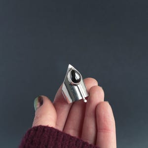 May include: A silver ring with a black teardrop-shaped gemstone. The ring is pointed and has a unique design.