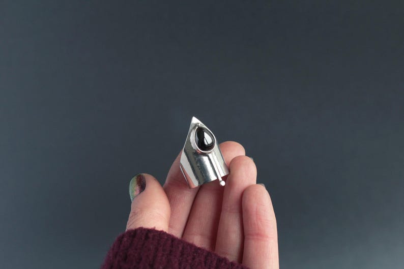 May include: A silver ring with a black teardrop-shaped gemstone. The ring is pointed and has a unique design.
