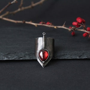 May include: A silver ring with a pointed design and a red gemstone in the center.