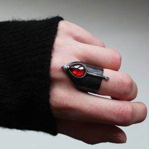 May include: A silver ring with a red gemstone set in a leaf-shaped design. The ring is worn on the index finger of a hand.