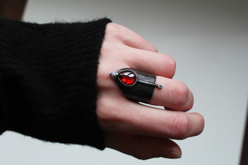 May include: A silver ring with a red gemstone set in a leaf-shaped design. The ring is worn on the index finger of a hand.