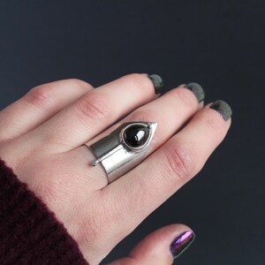 May include: A silver ring with a black gemstone set in a pointed, triangular design. The ring is worn on a finger with dark nail polish.