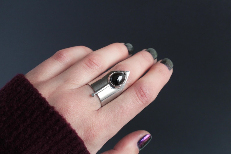 May include: A silver ring with a black gemstone set in a pointed, triangular design. The ring is worn on a finger with dark nail polish.