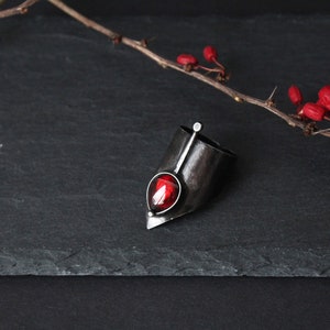 May include: A silver ring with a red gemstone set in a pointed, angular design. The ring is on a black slate surface with a branch of red berries.