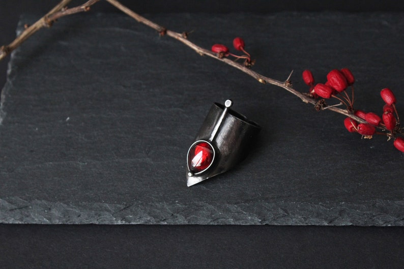 May include: A silver ring with a red gemstone set in a pointed, angular design. The ring is on a black slate surface with a branch of red berries.