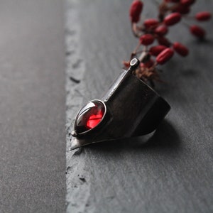 May include: A silver ring with a red gemstone set in a pointed, angular design. The ring is on a dark gray surface.