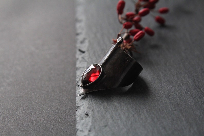 May include: A silver ring with a red gemstone set in a pointed, angular design. The ring is on a dark gray surface.