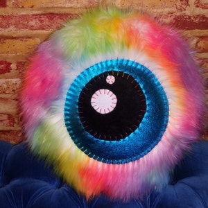 May include: A colorful, fluffy pillow with a large, black eye design. The eye is surrounded by a blue, sparkly circle and has a white pupil.