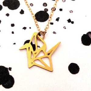 May include: A gold origami crane pendant necklace on a delicate gold chain. The necklace is photographed against a white background with black splatter paint.