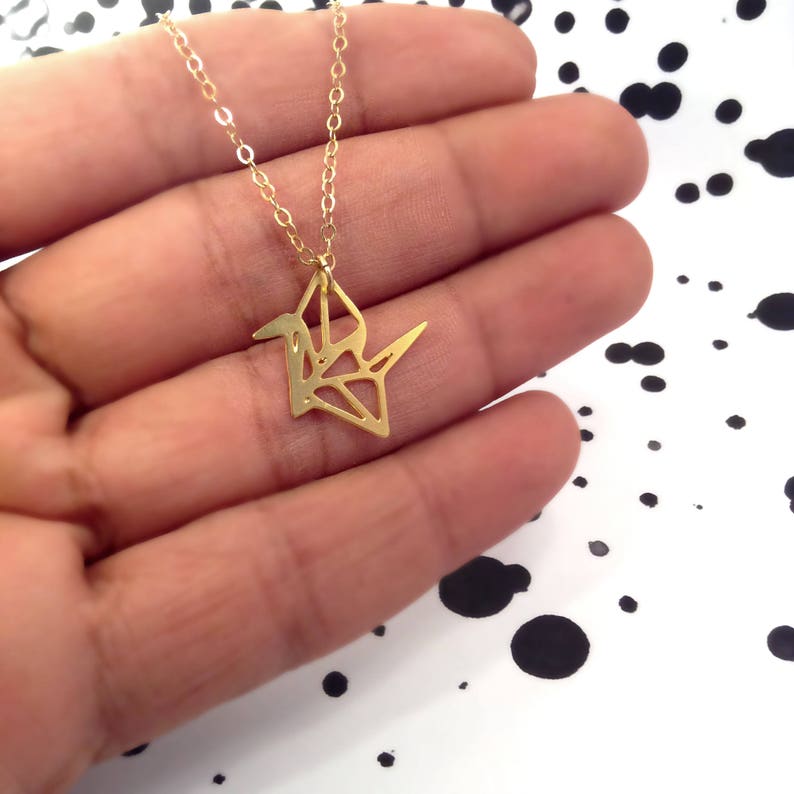 May include: A gold necklace with a delicate origami crane pendant. The crane is made of a single piece of metal and has a detailed, geometric design.