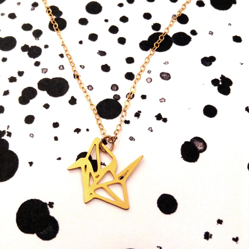 May include: A gold origami crane pendant necklace on a delicate gold chain. The necklace is photographed against a white background with black splatter paint.