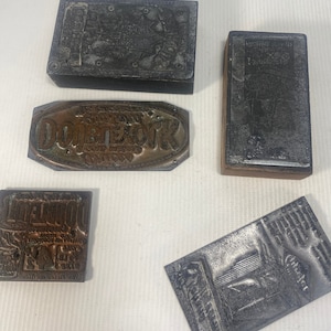 May include: Five vintage printing blocks, including a copper block with the text "The Original" and "DOBSTERK" in a circular design, a wooden block with a metal plate with the text "X-50", and three metal blocks with various designs.