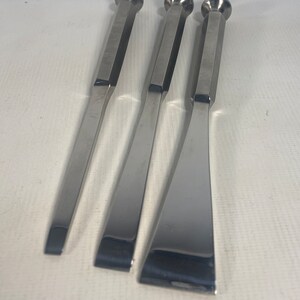 May include: Three silver metal tools with rounded ends and flat blades. The tools are arranged in a row, with the largest tool on the right and the smallest tool on the left.