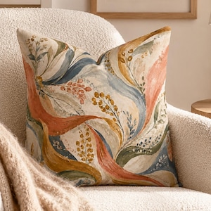 May include: A square throw pillow featuring a watercolor-style botanical pattern with flowing, abstract leaf and vine motifs in shades of terracotta orange, muted blue, sage green, and golden yellow on a light cream-colored fabric background.