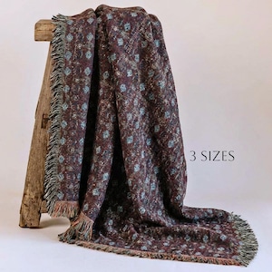 May include: A woven throw blanket featuring a dark maroon base with a repeating pattern of small, light blue diamond shapes and a fringed edge. The blanket is draped over a wooden stool and includes the text 3 SIZES printed on the right side.
