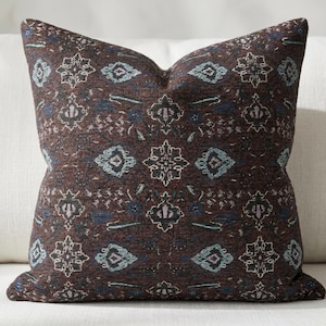 May include: A decorative throw pillow with a dark brown background and an intricate pattern in shades of blue, teal, and off-white. The pattern features geometric and floral elements, creating a textured appearance. The pillow is placed on a white surface.