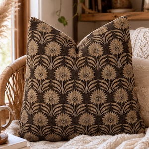 May include: A square throw pillow featuring a repeating pattern of tan sunflowers with dark brown stems and leaves set against a dark brown background. The pillow has a textured fabric appearance and is displayed in a home setting.