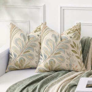 May include: Two square throw cushions featuring a watercolour-style botanical pattern with large, stylised blue and green leaves on a cream-coloured background. The design includes delicate stems and small dotted details throughout the foliage.