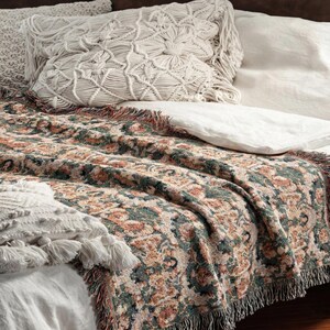 May include: A bed with a patterned throw blanket and decorative pillows. The throw blanket features a floral design in shades of green, orange, and beige, with a fringed edge. The pillows are white and cream-colored, with macrame and tassel details.