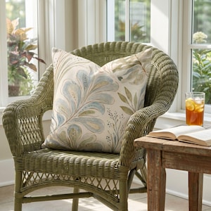 May include: A square throw cushion featuring a watercolour-style botanical print with large, stylised blue and green leaves on a cream background. The cushion is placed on a green woven wicker chair with a curved back and armrests.