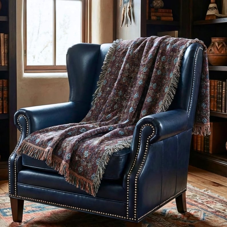 May include: A navy blue leather wingback chair with a patterned throw blanket. The blanket has a burgundy background with blue and tan floral designs and fringe. The chair has brass nailhead trim.